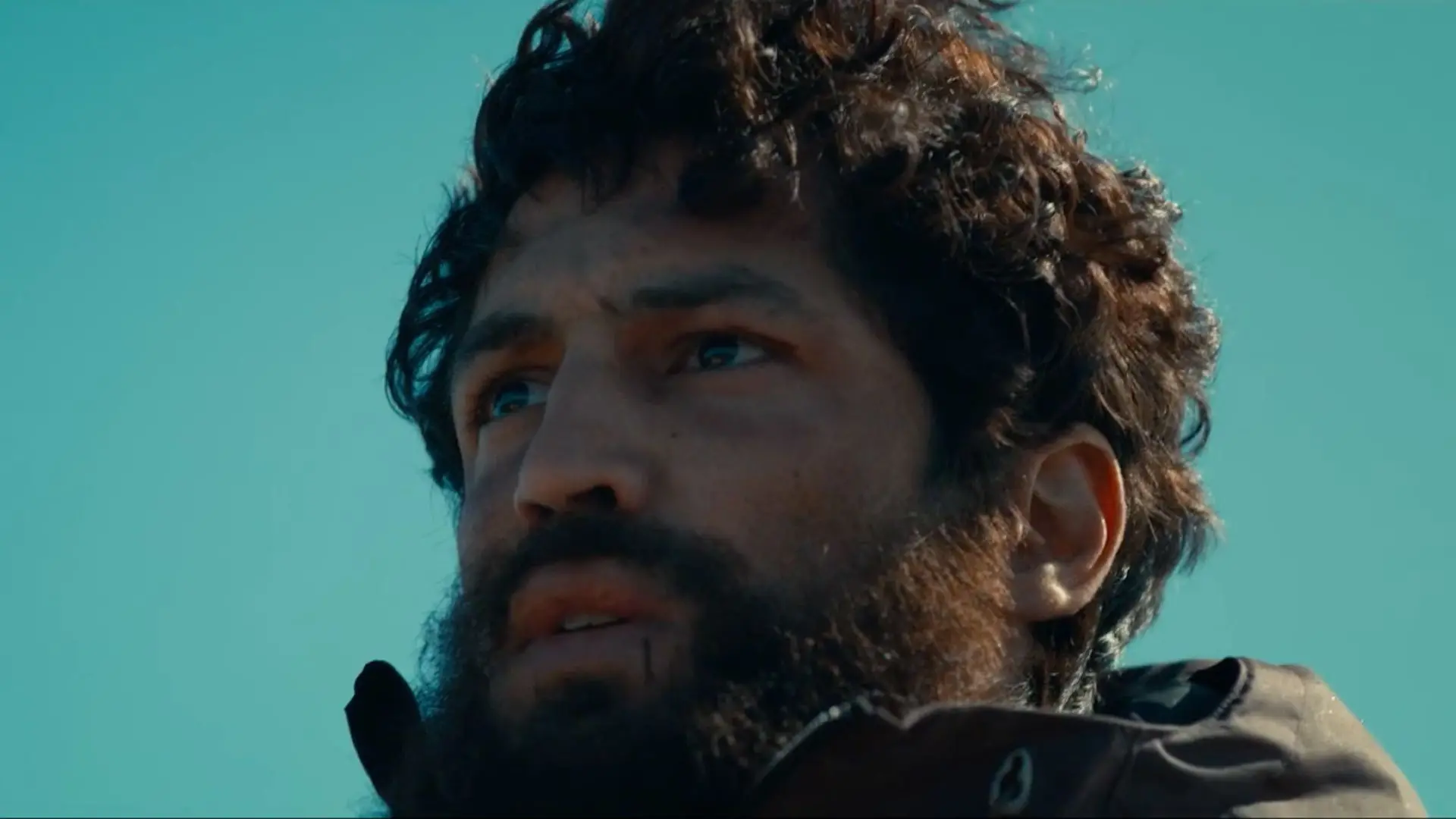 Pessoa ao ar livre, em close, com barba espessa e cabelo curto e ondulado, usando casaco escuro; ao fundo, céu azul.