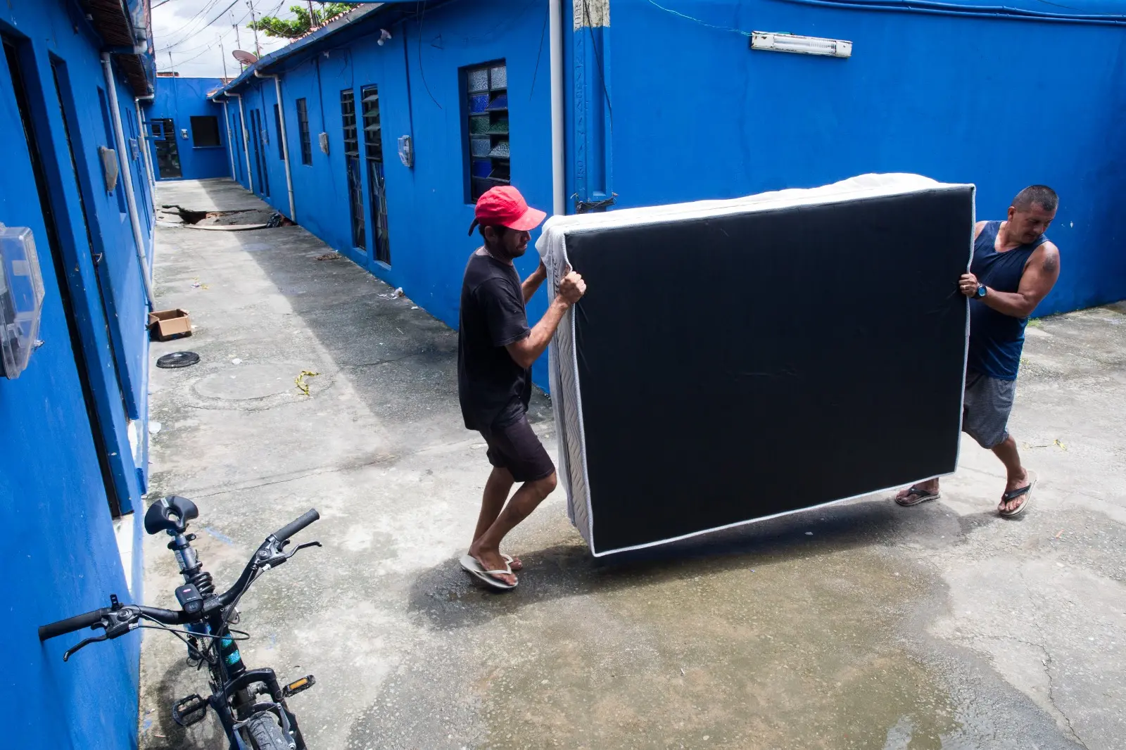 Dois homens seguram colchão em mudança de vila com paredes azuis onde chão afundou em Fortaleza.