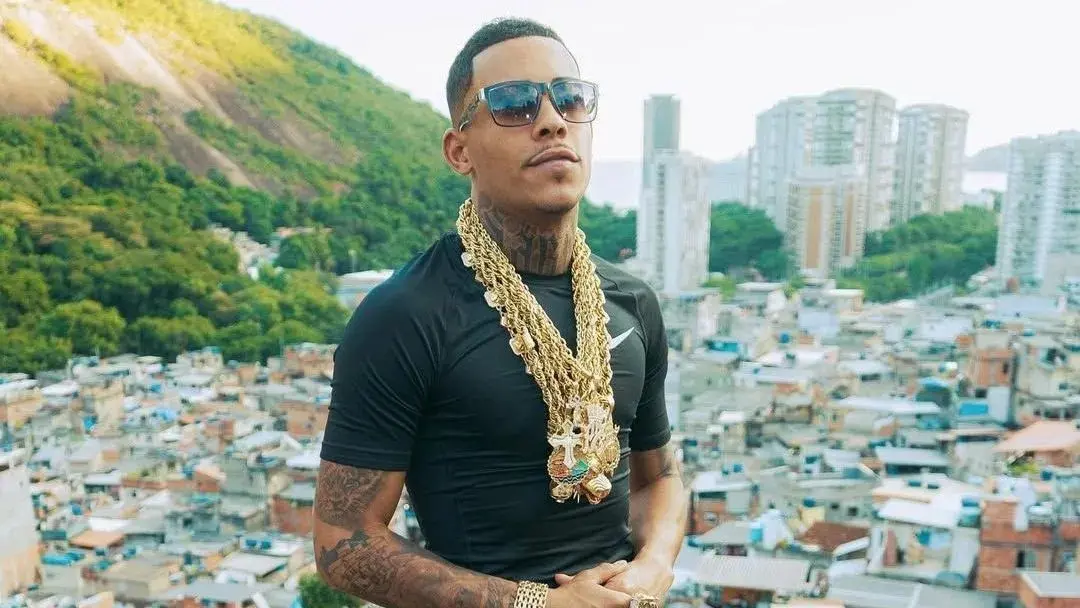 MC Poze do Rodo posa usando óculos escuros e diversas correntes de ouro grossas sobre uma camiseta preta em um mirante. Ao fundo, a paisagem destaca o contraste entre o verde das montanhas e a densa urbanização de uma comunidade, sob a luz clara do dia.