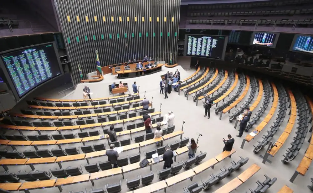Vista ampla do plenário de uma casa legislativa, com fileiras curvas de assentos quase vazios, poucos parlamentares e assessores dispersos, e painel eletrônico com votações ao fundo.