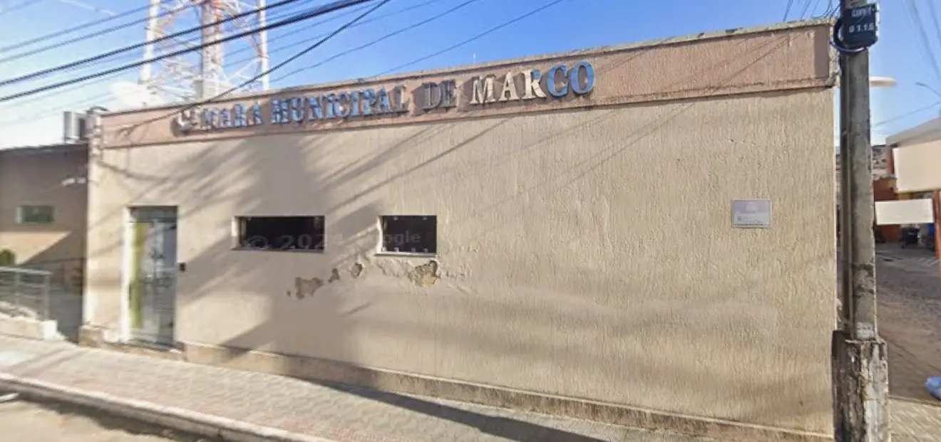 Imagem de uma fachada simples de prédio público com a inscrição “Câmara Municipal de Marco” na parte superior. O edifício tem paredes em tom bege, com sinais de desgaste e pequenas áreas descascadas. Há uma porta à esquerda e duas janelas estreitas com grades. A rua em frente é calçada, com fios elétricos visíveis acima e postes ao redor. O céu está azul e ensolarado, indicando um dia claro.