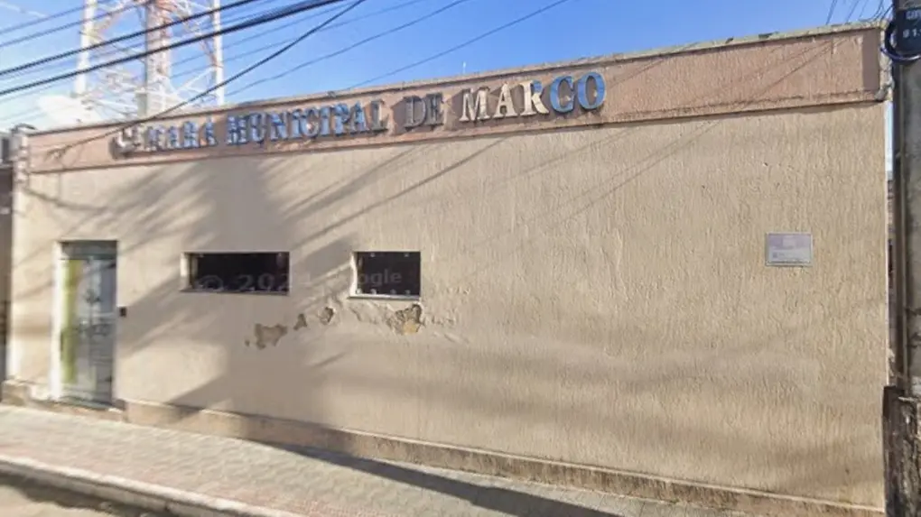 Imagem de uma fachada simples de prédio público com a inscrição “Câmara Municipal de Marco” na parte superior. O edifício tem paredes em tom bege, com sinais de desgaste e pequenas áreas descascadas. Há uma porta à esquerda e duas janelas estreitas com grades. A rua em frente é calçada, com fios elétricos visíveis acima e postes ao redor. O céu está azul e ensolarado, indicando um dia claro.