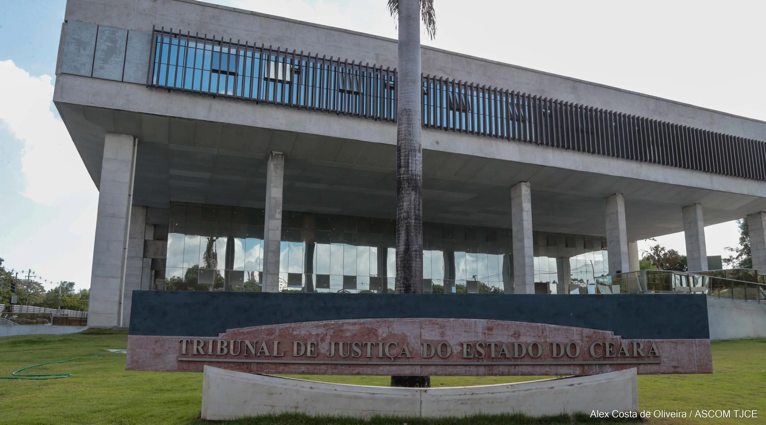 Fachada do Tribunal de Justiça do Estado do Ceará (TJCE), em Fortaleza. O prédio moderno é elevado sobre pilares de concreto, com grandes janelas de vidro refletindo o céu. Em primeiro plano, há um jardim gramado e um letreiro com a inscrição TRIBUNAL DE JUSTIÇA DO ESTADO DO CEARÁ
