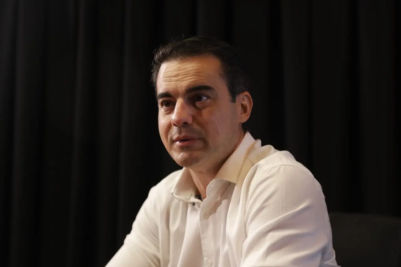 Homem de meia-idade, com cabelos escuros e curtos, usando camisa branca, aparece em close médio contra um fundo escuro de cortina. Ele está levemente virado para a direita, com expressão atenta e séria, como se estivesse participando de uma entrevista ou conversa. A iluminação suave destaca seu rosto, criando contraste com o fundo preto.