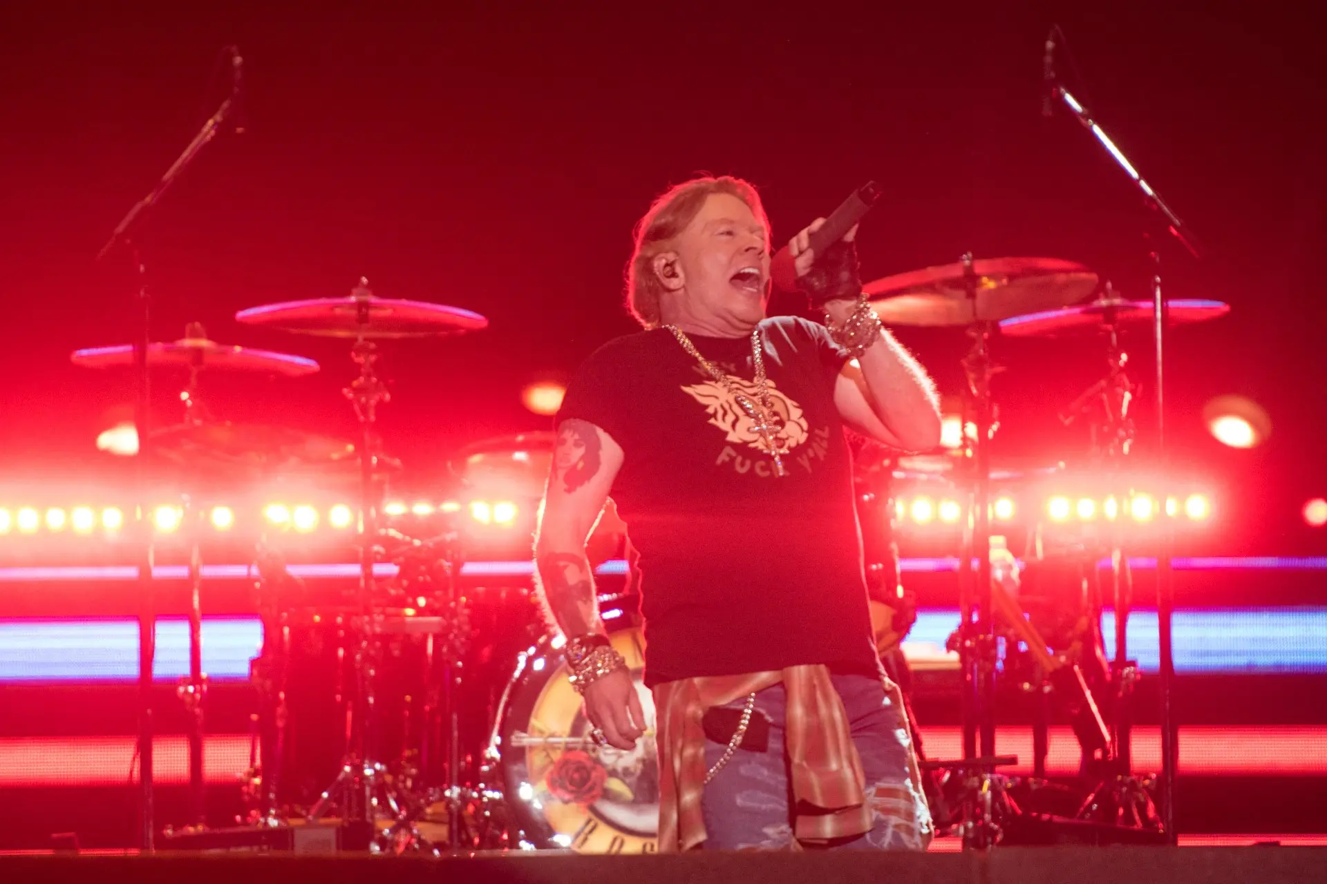 Axl Rose, da banda Guns N' Roses, se apresenta ao vivo em um palco com iluminação intensa, tocando guitarra e cantando em um show de rock.