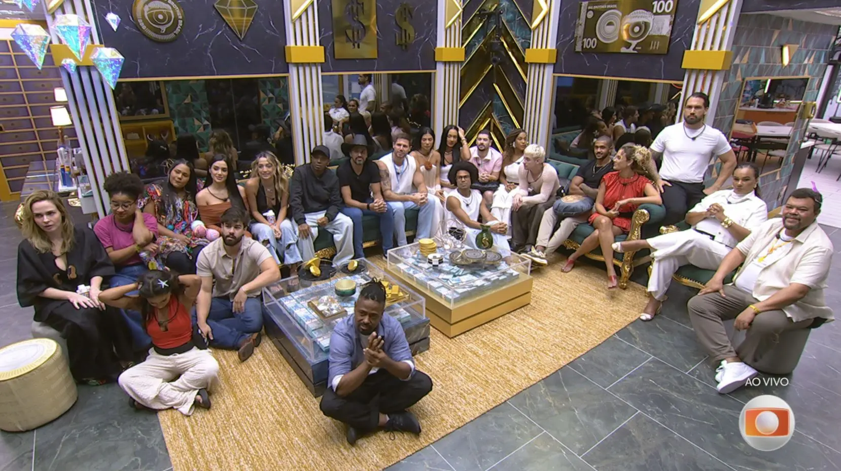 Sala de estar de um reality show com cerca de vinte participantes sentados em sofás e no chão, dispostos em semicírculo ao redor de mesas de centro de vidro, em ambiente interno decorado, com tapete claro e logotipo “AO VIVO” no canto da tela.