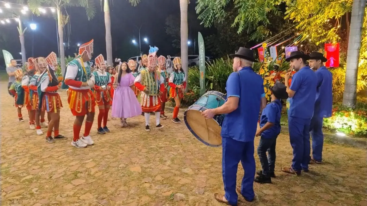 Grupo de pessoas participa de apresentação cultural ao ar livre, à noite. À esquerda, dançarinos com trajes coloridos e máscaras formam uma roda. Ao centro, uma pessoa com vestido lilás acompanha a dança. À direita, músicos vestindo azul tocam instrumentos de percussão. O local é iluminado, com árvores e decoração ao fundo.