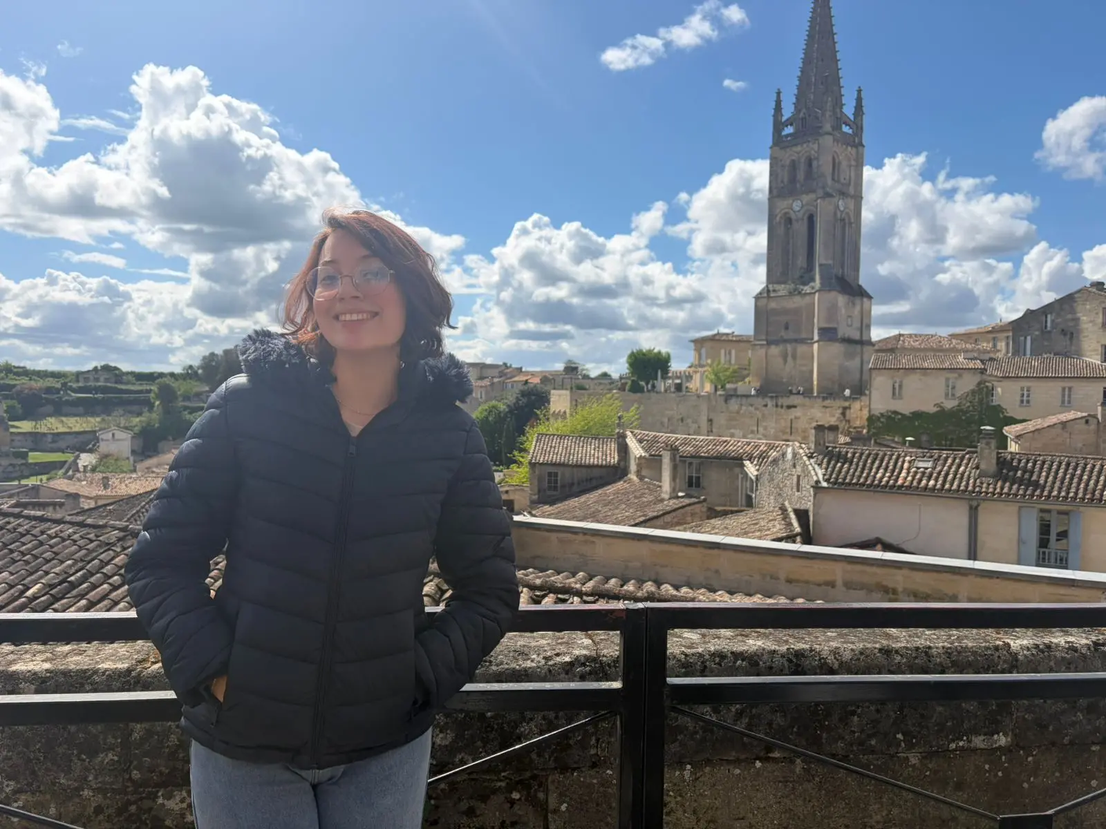 Pessoa sorridente, usando óculos e casaco escuro, está apoiada em uma grade com as mãos nos bolsos, em um mirante. Ao fundo, há telhados de casas de uma cidade histórica e uma igreja com torre alta sob céu azul com nuvens.