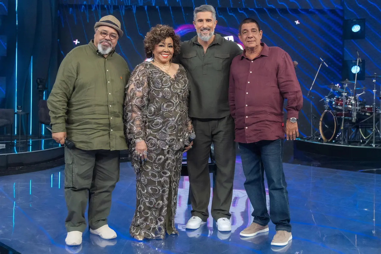 Jorge Aragão, Alcione, Marcos Mion e Zeca Pagodinho sorriem juntos no palco do programa 'Caldeirão'. O cenário vibrante apresenta tons de azul, luzes de LED ao fundo e instrumentos musicais dispostos na lateral, capturando um encontro festivo entre ícones da música.