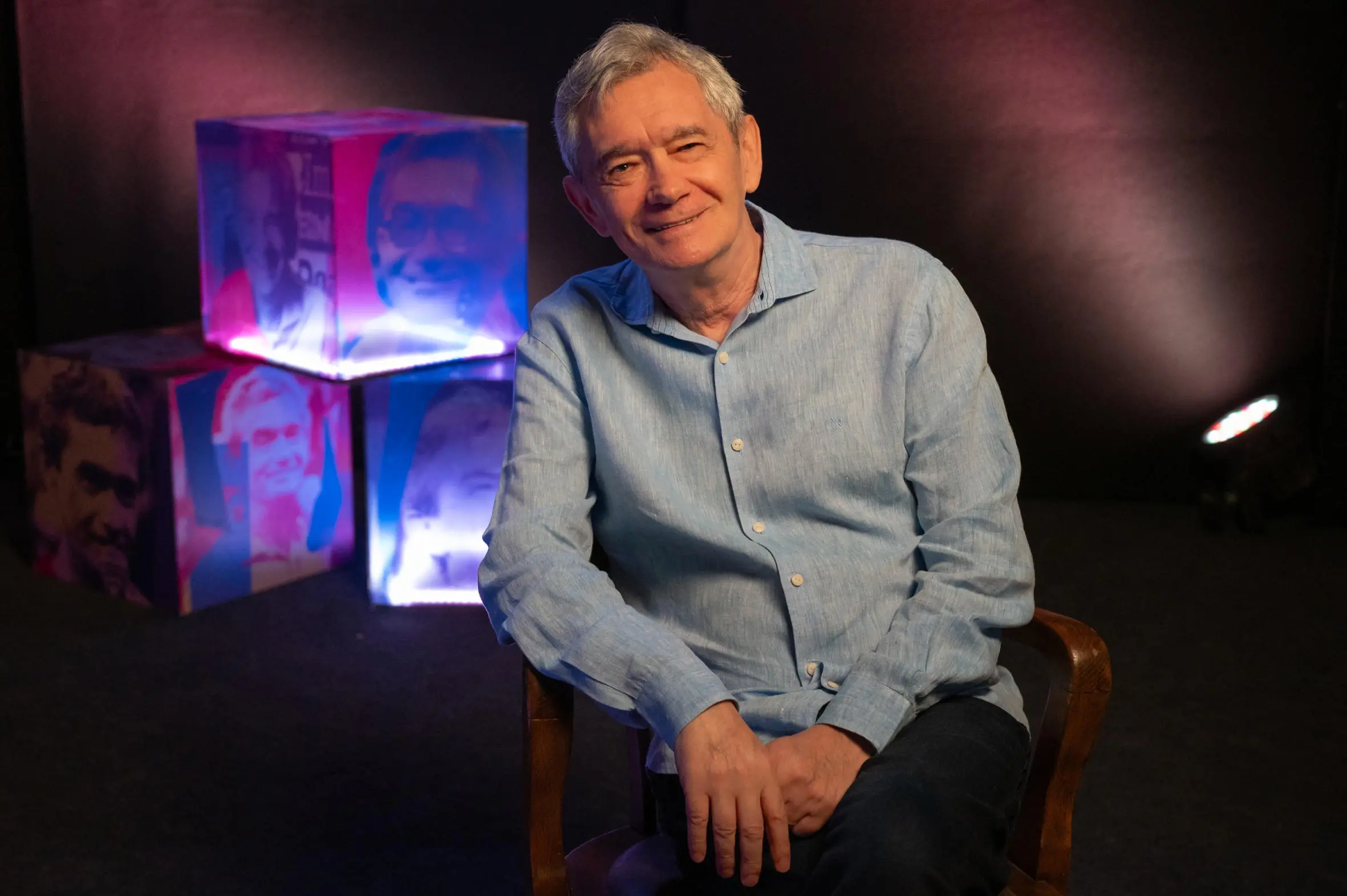 Serginho Groisman sorri sentado em uma cadeira de madeira, vestindo uma camisa de linho azul clara em um ambiente de estúdio com iluminação suave. Ao fundo, cubos iluminados exibem fotografias de seu rosto em tons de rosa e azul, criando uma composição moderna e nostálgica.