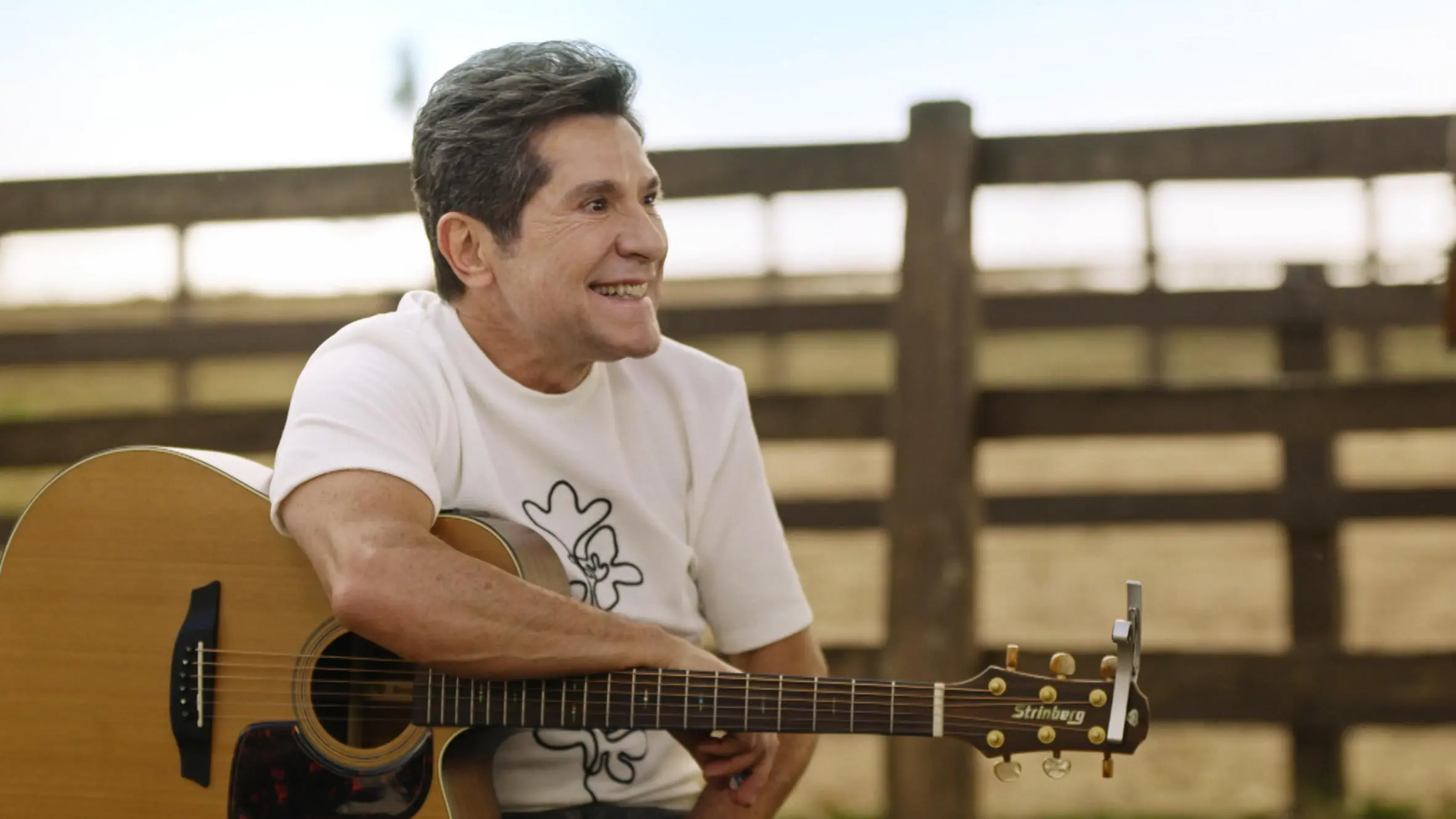 O cantor Daniel sorri enquanto segura seu violão, vestindo uma camiseta branca em um ambiente rural com uma cerca de madeira ao fundo. A cena é iluminada pela luz natural do dia, transmitindo uma atmosfera tranquila e rústica que remete ao universo sertanejo.