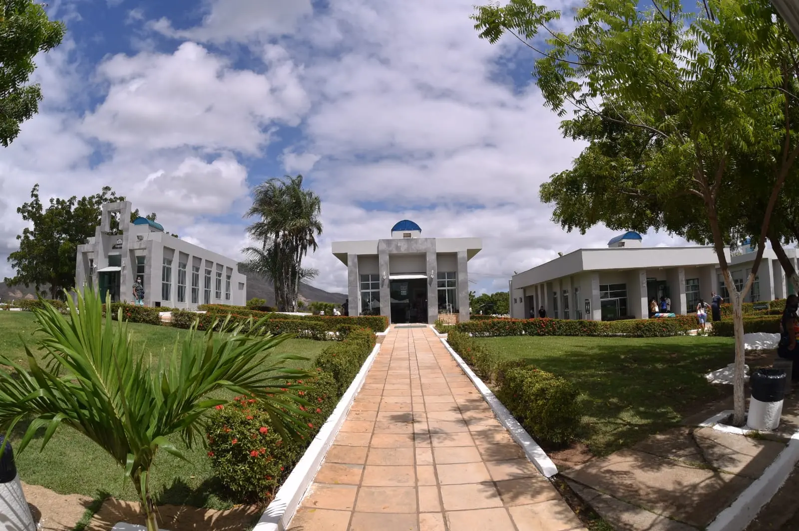 Uma passarela de pedra clara leva a modernos prédios cinzas com cúpulas azuis, cercados por gramados verdes e arbustos sob um céu ensolarado. Localizado em Maracanaú, o Memorial da Paz apresenta arquitetura contemporânea integrada à natureza em um ambiente sereno e arborizado.