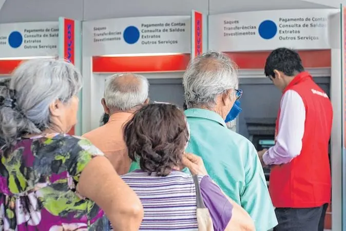 Fila de quatro idosos de costas, aguardando para usar um caixa eletrônico de banco com o auxílio de um funcionário da instituição.