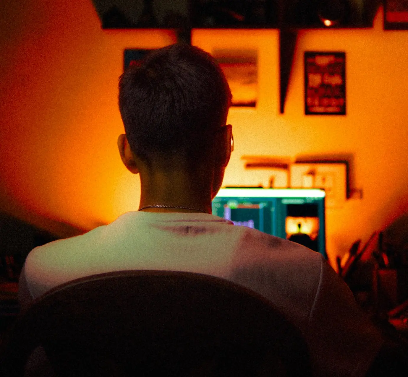 Vista de costas de um jovem sentado em frente a um computador em um ambiente com iluminação quente e amarelada. O monitor exibe uma interface de edição, enquanto a parede ao fundo decorada com quadros completa a atmosfera de trabalho criativo.