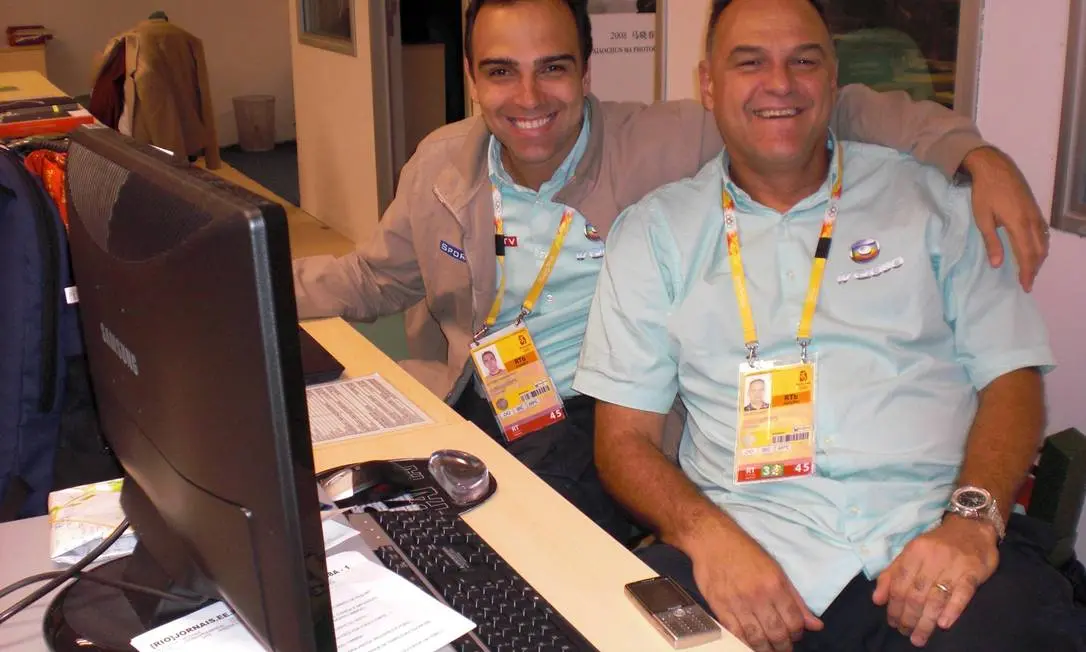 Tadeu Schmidt e Oscar Schmidt sorriem abraçados em uma bancada de trabalho durante a cobertura das Olimpíadas de Pequim, ambos usando credenciais oficiais e camisas de uniforme. O ambiente de redação ao fundo conta com computadores e equipamentos de transmissão, registrando um momento de descontração entre os irmãos.