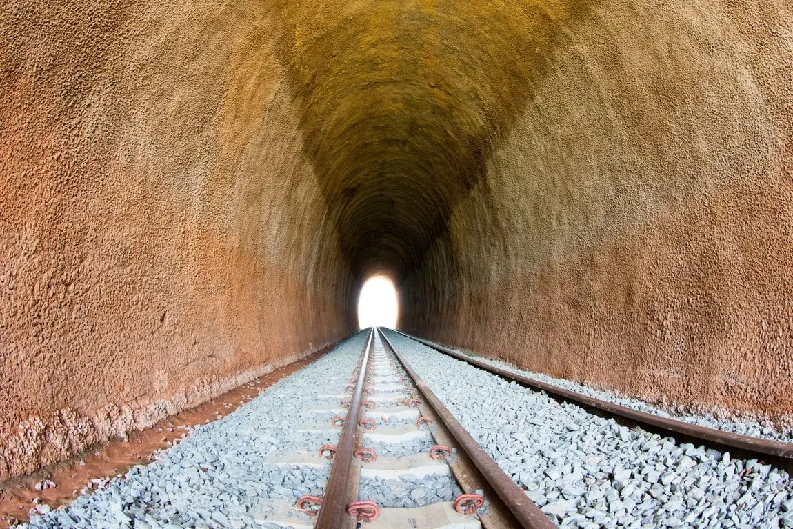 Foto que contém trilho ferroviário novo, ao fundo com luz no fim do túnel.