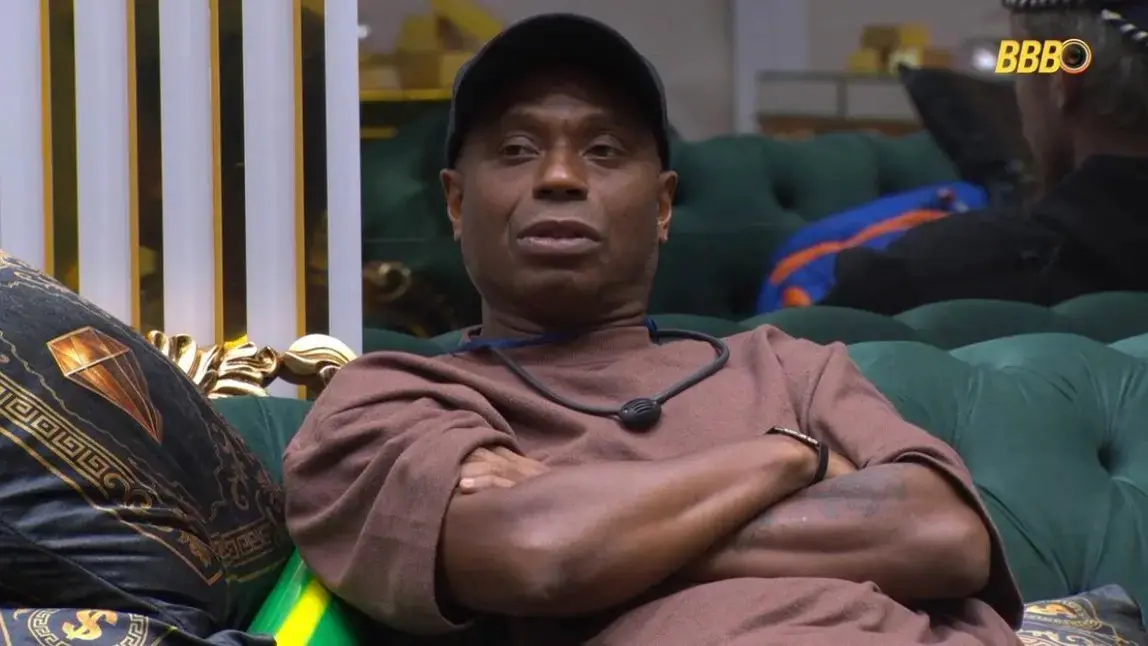 Edilson é um homem idoso e negro. Na foto, ele usa camisa marrom e boné preto e está com os braços cruzados.