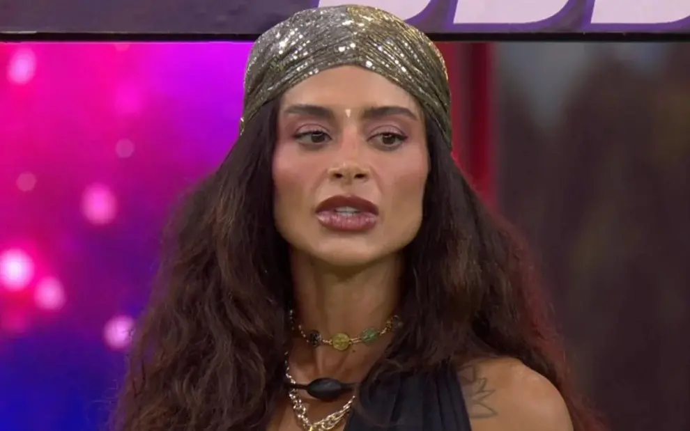 Aline Campos é uma mulher de meia idade, branca, de cabelo longo e cheio, castanho. Na foto, ela usa uma touca brilhosa e acessórios também brilhosos.