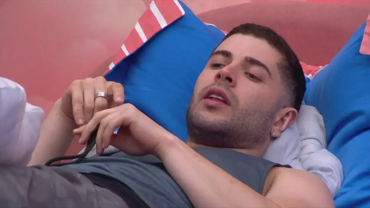 Marcelo é um homem jovem, branco e de barba rala. Na foto, ele está deitado na cama e usa camiseta regata.