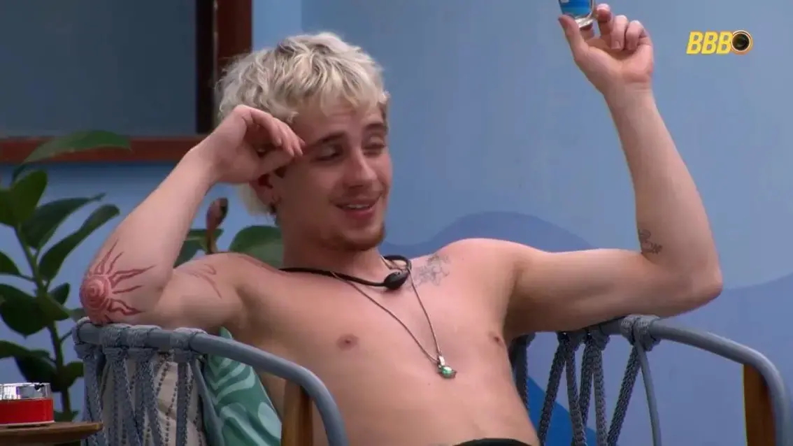 Juliano Floss é um homem jovem, branco, de cabelo loiro descolorido. Na foto, está sem camisa, dialogando com alguém.