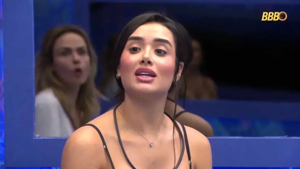 Jordana é uma mulher jovem branca de cabelo preto amarrado em um coque. Na foto, ela está de blusa de alça dialogando com alguém.