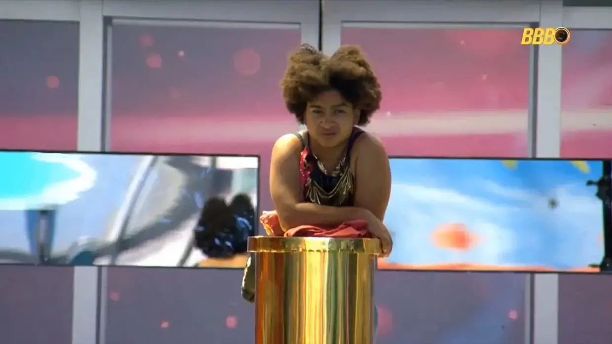 Milena é uma jovem negra de cabelo black. Na foto, ela está apoiada no totem dourado do Castigo do Monstro.