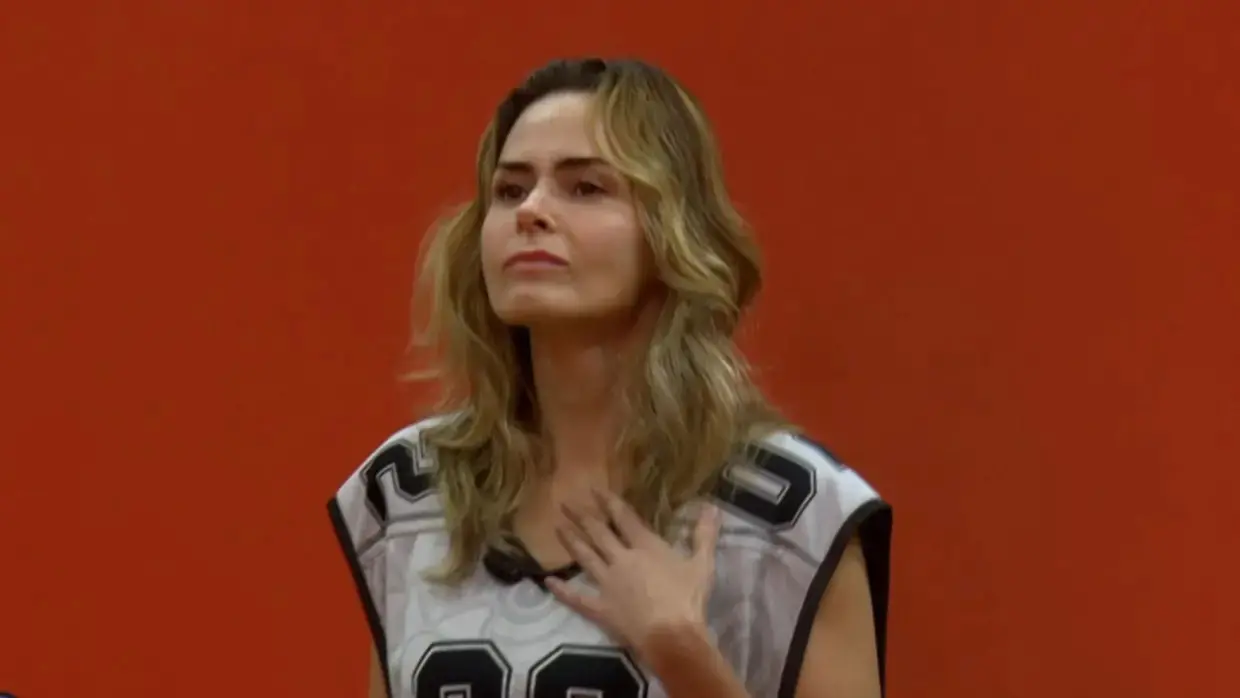 Ana Paula é uma mulher branca de meia idade, loira e de cabelo ondulado. Na foto, ela usa colete de uma das provas do BBB.