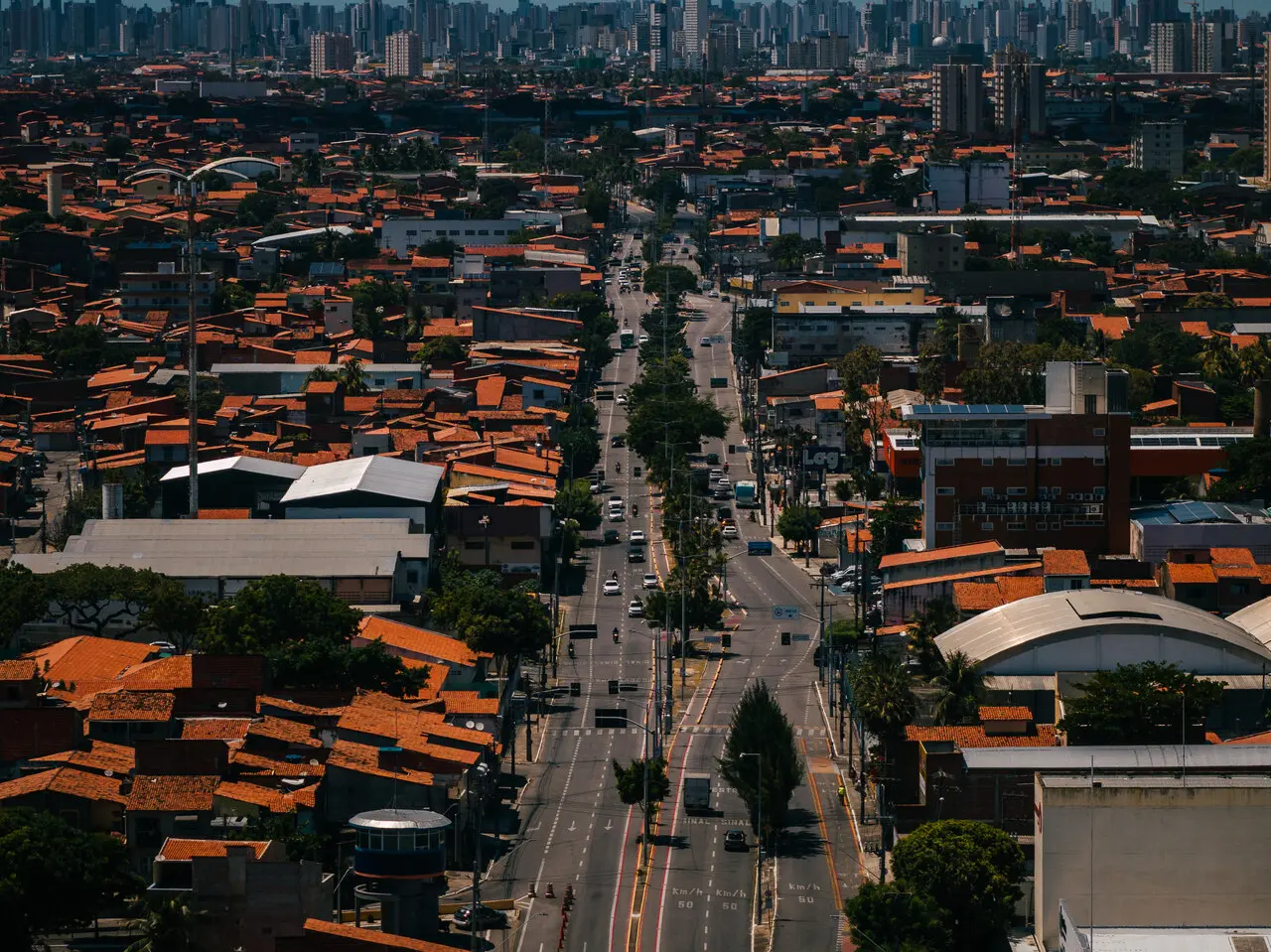 Uma avenida movimentada corta o bairro da Barra do Ceará, com o fluxo de veículos serpenteando entre casas de telhados cerâmicos alaranjados e galpões industriais. Ao fundo, a densa linha de prédios modernos no horizonte contrasta com a arquitetura horizontal da região em uma perspectiva urbana profunda sob a luz do dia.