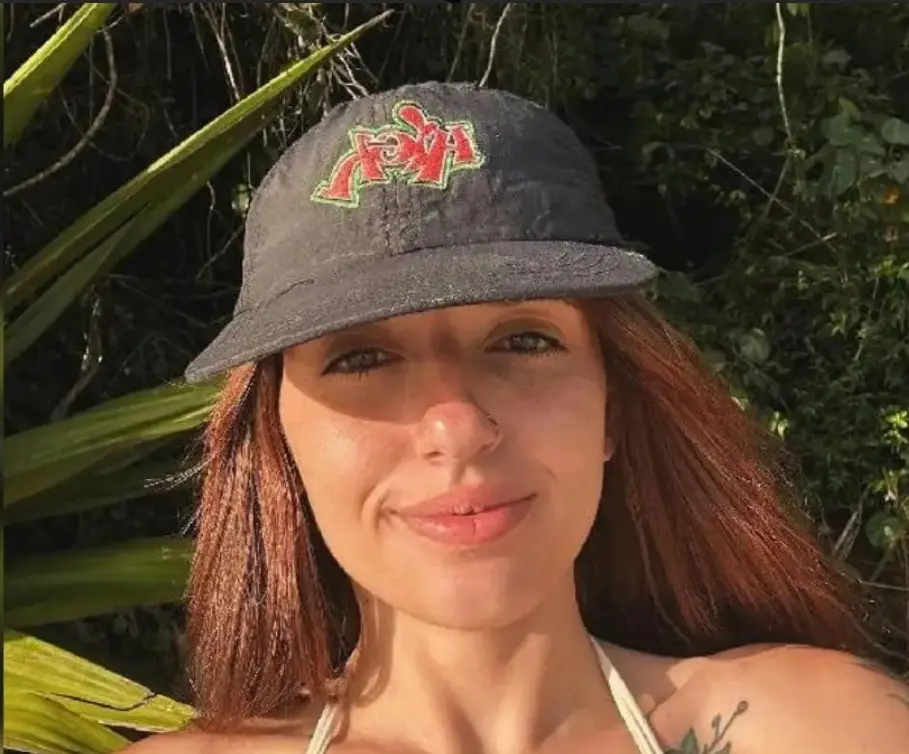 Uma mulher jovem de pele clara e longos cabelos castanhos-avermelhados sorri suavemente para a câmera, usando um boné preto com um bordado vermelho e verde. Ela veste um top branco de alças finas e exibe um piercing discreto no nariz, além de uma tatuagem de folhagens no ombro. A iluminação solar intensa cria sombras em seu rosto e destaca a vegetação verde e tropical ao fundo.