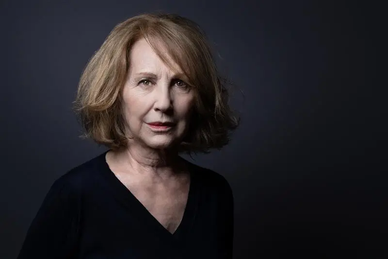 Um retrato de estúdio em plano médio mostra uma mulher madura de pele clara e expressão serena, com cabelos castanhos curtos e levemente ondulados. Ela veste uma blusa preta de decote em 