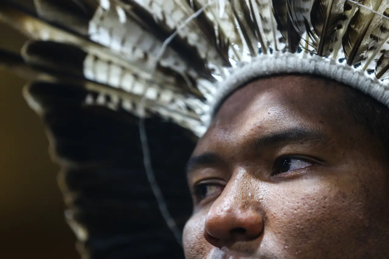 Close-up lateral do rosto de um homem indígena focado, destacando os detalhes de seu cocar feito de penas claras e escuras. A iluminação enfatiza a textura da pele e a expressão atenta do olhar.