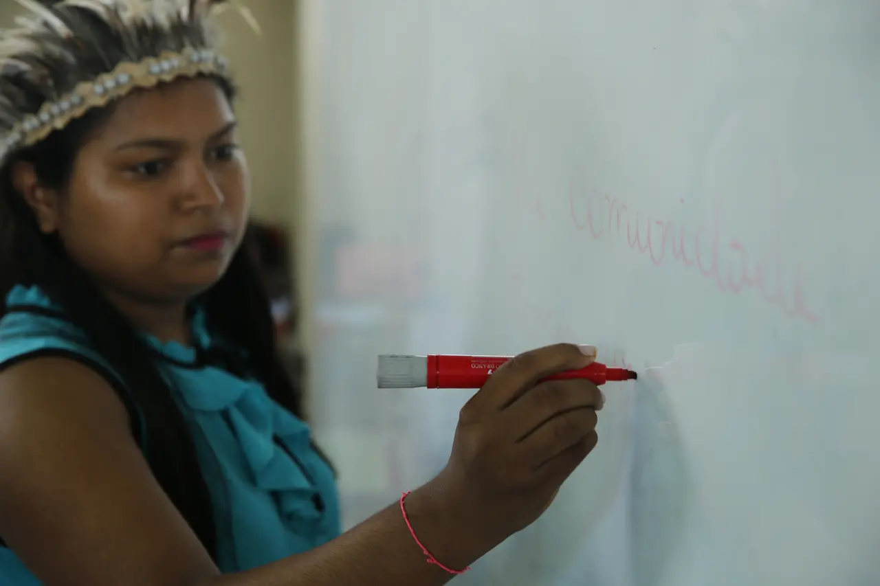 Uma mulher indígena de cocar escreve com um pincel atômico vermelho em um quadro branco. A imagem possui um foco seletivo na mão que escreve, mantendo o rosto da professora levemente desfocado ao fundo.