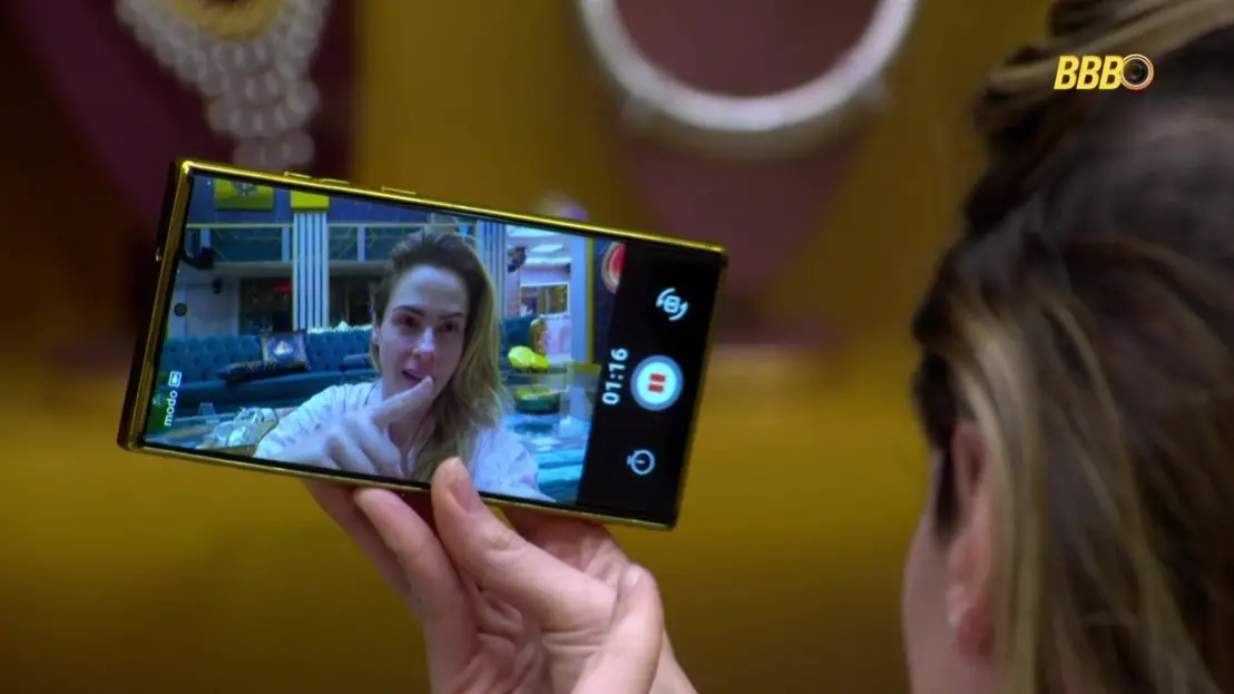 Close-up da tela de um smartphone mostrando Ana Paula gravando um depoimento. Ela aponta o dedo indicador para a câmera, usando um pijama rosa claro, com o cenário da sala da casa ao fundo.