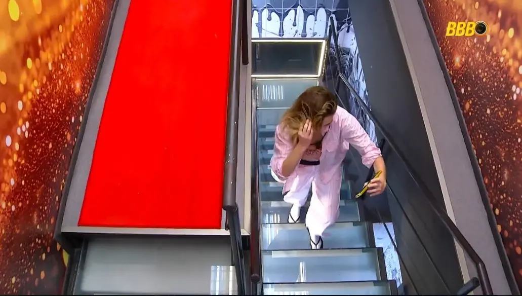 Vista aérea de uma participante de pijama rosa subindo uma escada de vidro enquanto grava um vídeo com o celular. Ao lado, há uma rampa com tapete vermelho e paredes decoradas com luzes douradas e artes gráficas pretas.