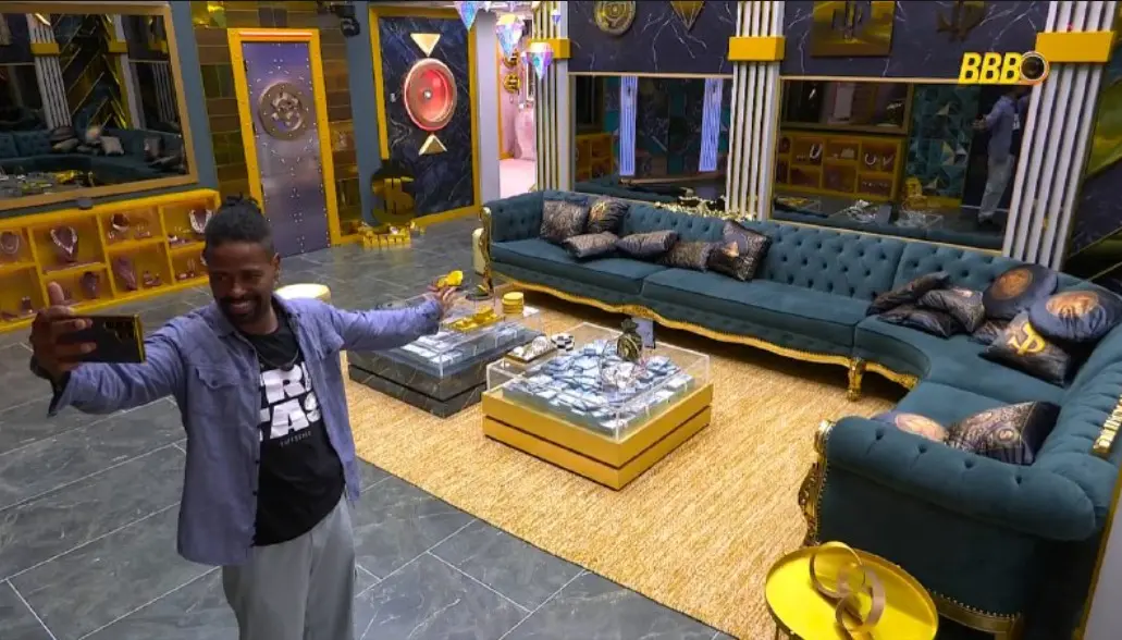 Leandro Boneco sorridente tira uma selfie na sala da casa, que tem decoração luxuosa em tons de azul e dourado. O ambiente conta com um grande sofá de veludo, tapete sisal e mesas de centro repletas de maços de dinheiro cenográfico.