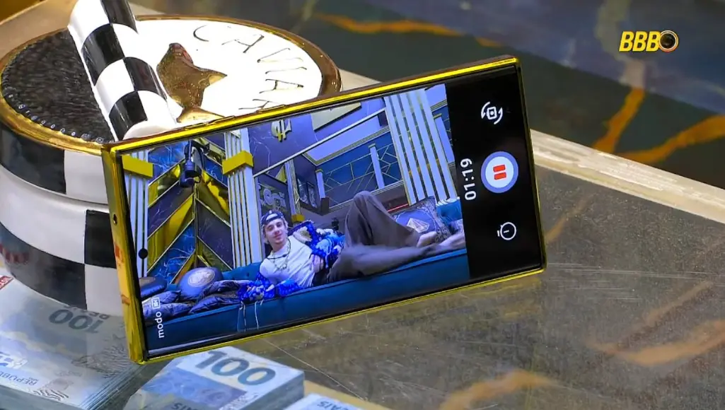 Um celular apoiado sobre uma mesa de vidro exibe a gravação de Juliano Floss de boné e jaqueta azul deitado no sofá da sala. Ao redor do aparelho, veem-se detalhes da decoração, como pilhas de notas de 100 reais cenográficas.