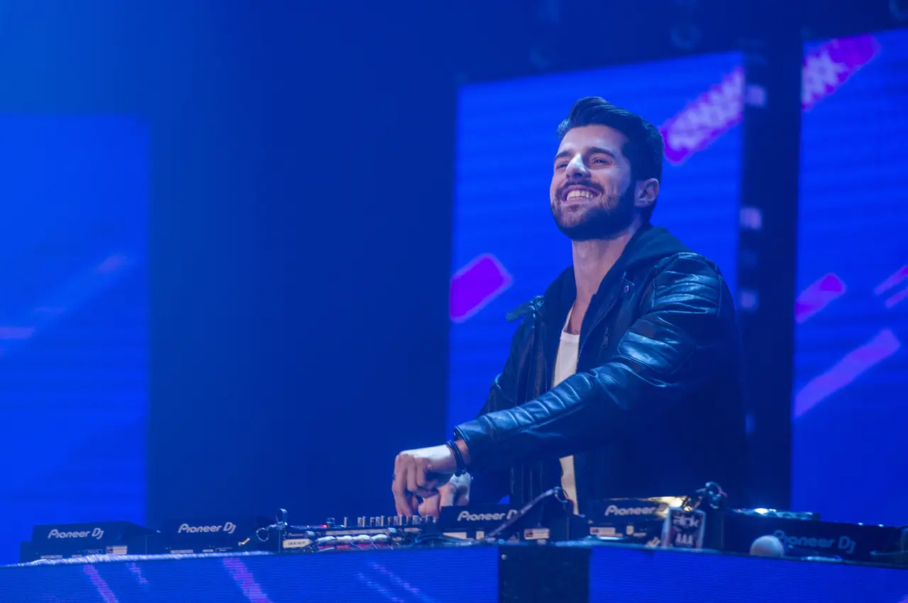 O DJ Alok sorri enquanto comanda sua mesa de som em um palco iluminado por intensos tons de azul e roxo. Ele veste uma jaqueta de couro preta sobre uma camiseta branca, posicionado em frente a grandes painéis de LED que exibem gráficos abstratos e vibrantes durante sua performance.