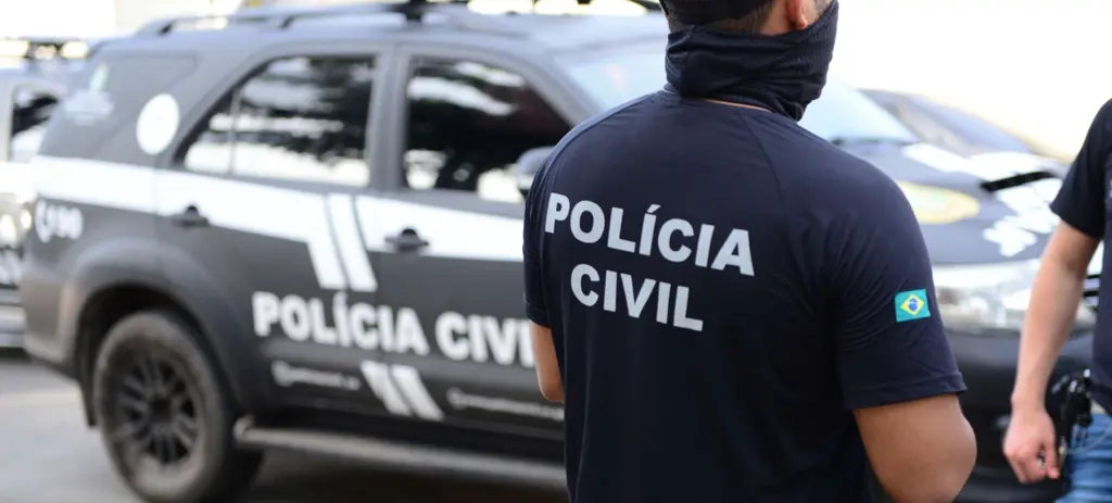 Agente da Polícia Civil visto de costas, usando camiseta com a inscrição “Polícia Civil”, em frente a uma viatura estacionada.