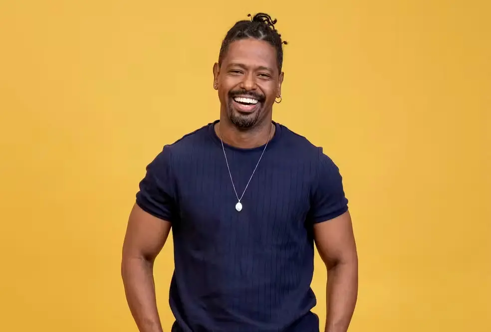 Leandro Boneco posa para foto em um fundo amarelo. Ele sorri para a câmera.