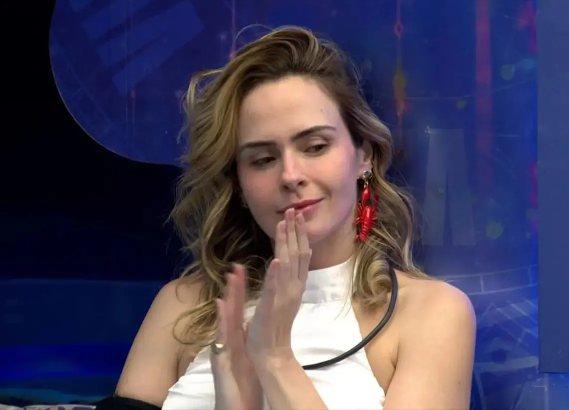 Pessoa sentada, com cabelo molhado, segura uma almofada estampada enquanto conversa com outra pessoa em ambiente interno com fundo azul pontilhado por pequenas luzes.