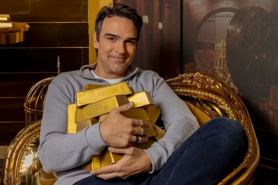 Homem sorridente segurando várias barras de ouro e sentando-se em uma cadeira dourada elegante, transmitindo sucesso e riqueza.