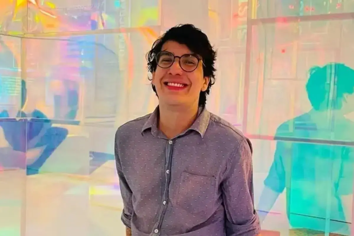 Imagem de rede social do professor brasileiro Danilo Neves Pereira, encontrado morto na Argentina. No registro, ele sorri para a câmera, usando óculos de armação redonda e uma camisa social cinza de mangas dobradas. Ele está posicionado à frente de uma estrutura de painéis translúcidos e coloridos que refletem tons vibrantes de rosa, azul e amarelo.