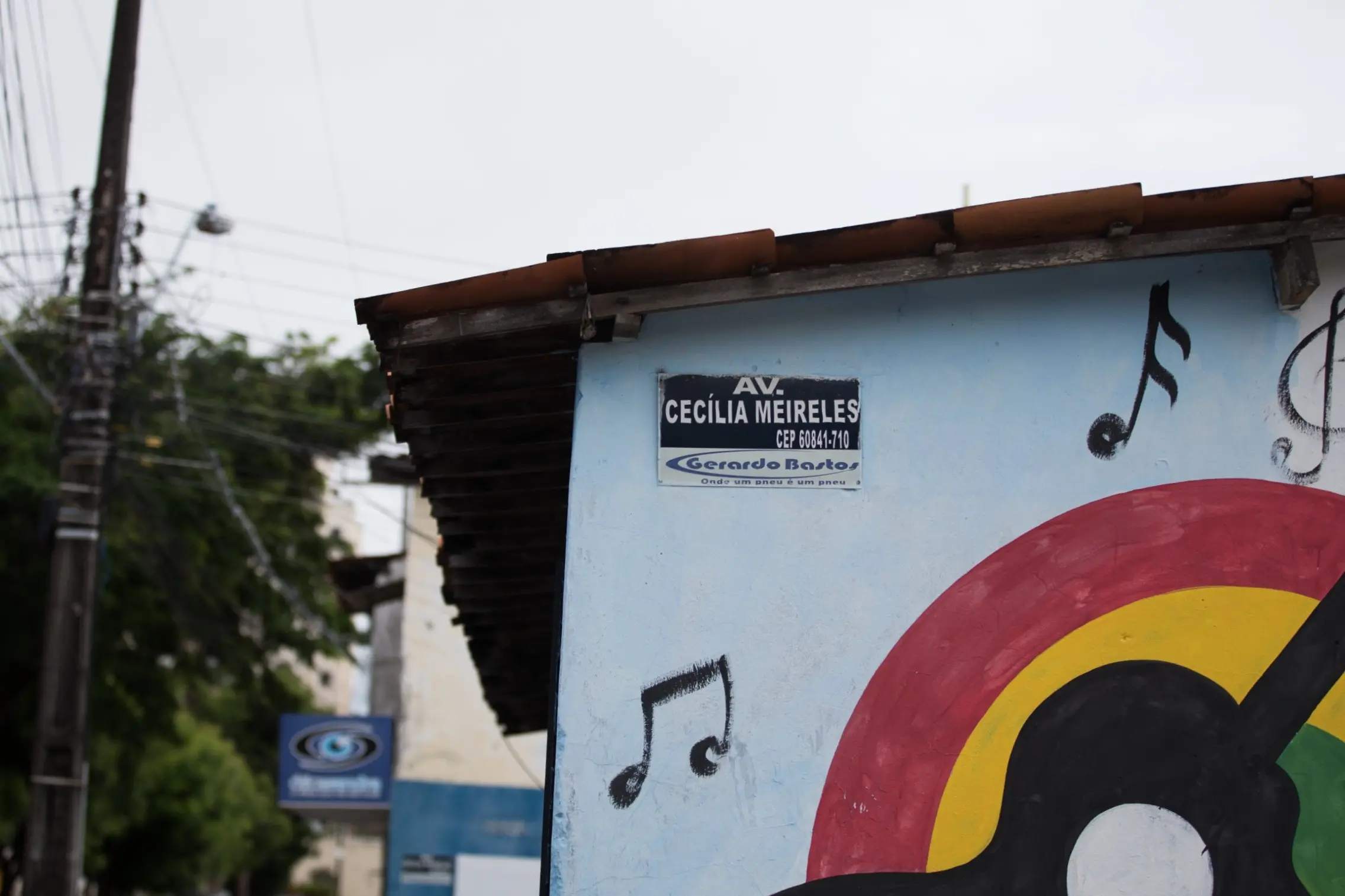 Placa da Avenida Cecília Meireles em parede azul com grafite de notas musicais. Ao fundo, vista de rua arborizada com poste de energia elétrica e céu nublado.