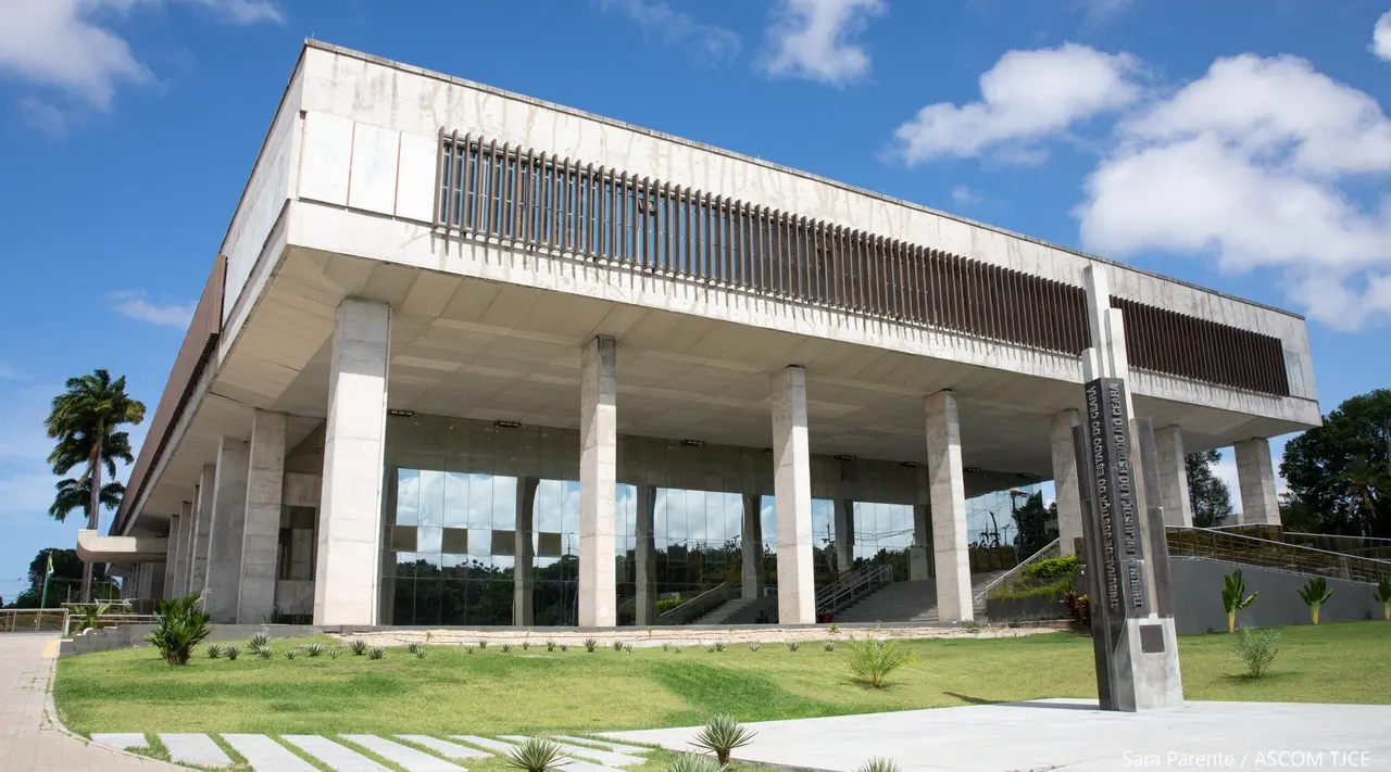 Fachada do Tribunal de Justiça do Ceará, edifício moderno de concreto e vidro com colunas altas, fachada retangular suspensa, escadaria frontal, jardim gramado e céu azul com nuvens ao fundo, em Fortaleza.