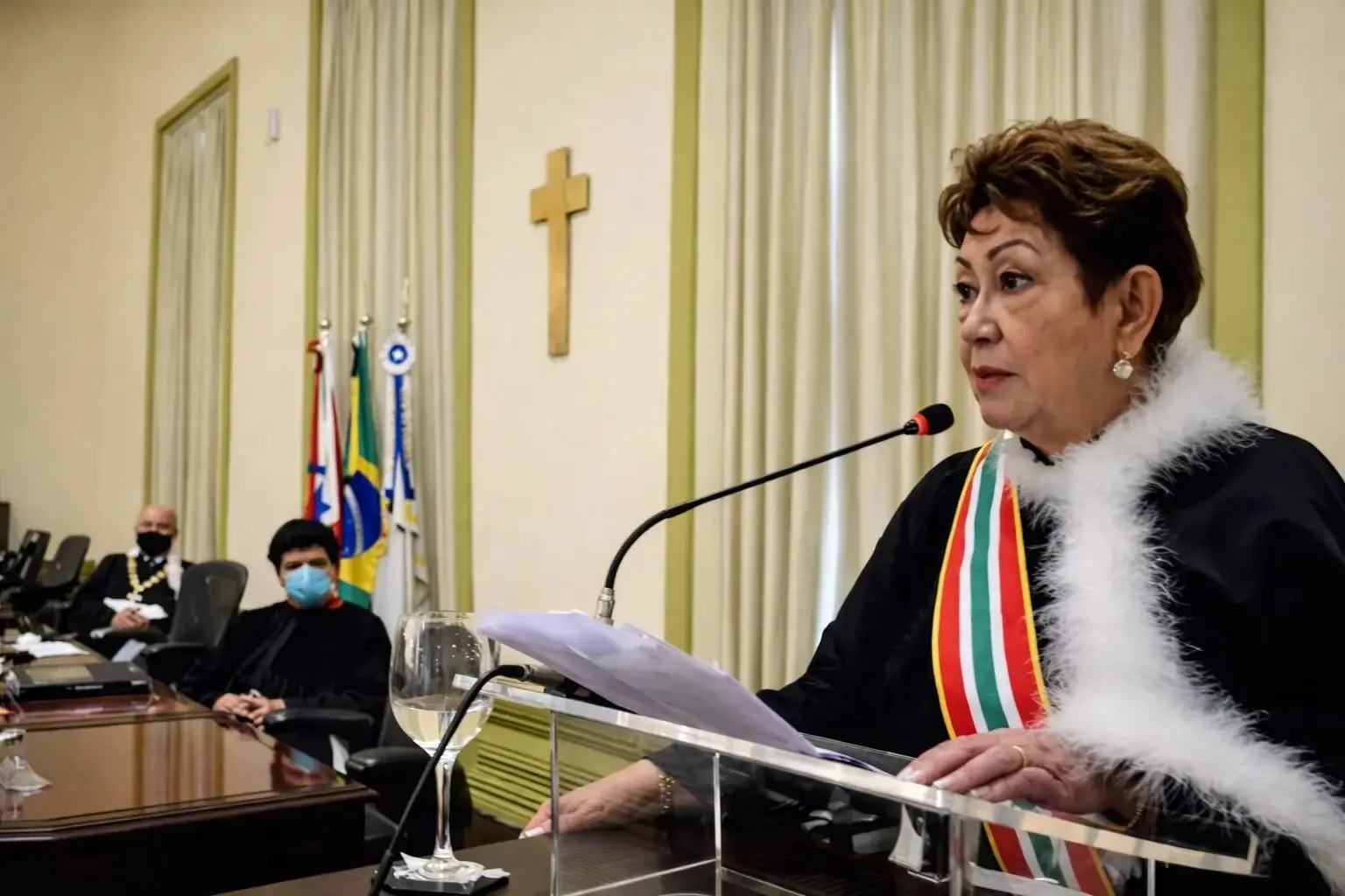 Desembargadora Eva do Amaral Coelho discursa ao microfone em sessão solene, atrás de um púlpito transparente, usando traje cerimonial com faixa tricolor. Ao fundo, integrantes da mesa diretora acompanham a cerimônia sentados, alguns com máscara facial, em uma sala institucional com bandeiras oficiais e um crucifixo na parede.
