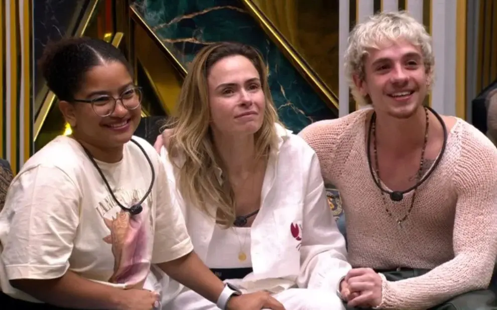 Milena, Ana Paula e Juliano sentados no sofá da sala do BBB 26, com expressão emotiva.