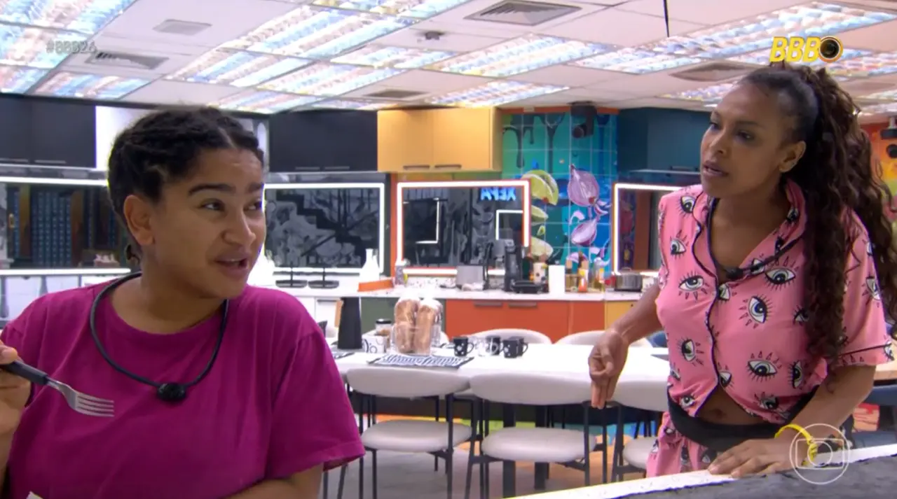 Duas pessoas em pé na cozinha de uma casa de reality show, uma segurando um garfo próximo à mesa e outra gesticulando; ao fundo, mesas, bancos e decoração colorida.