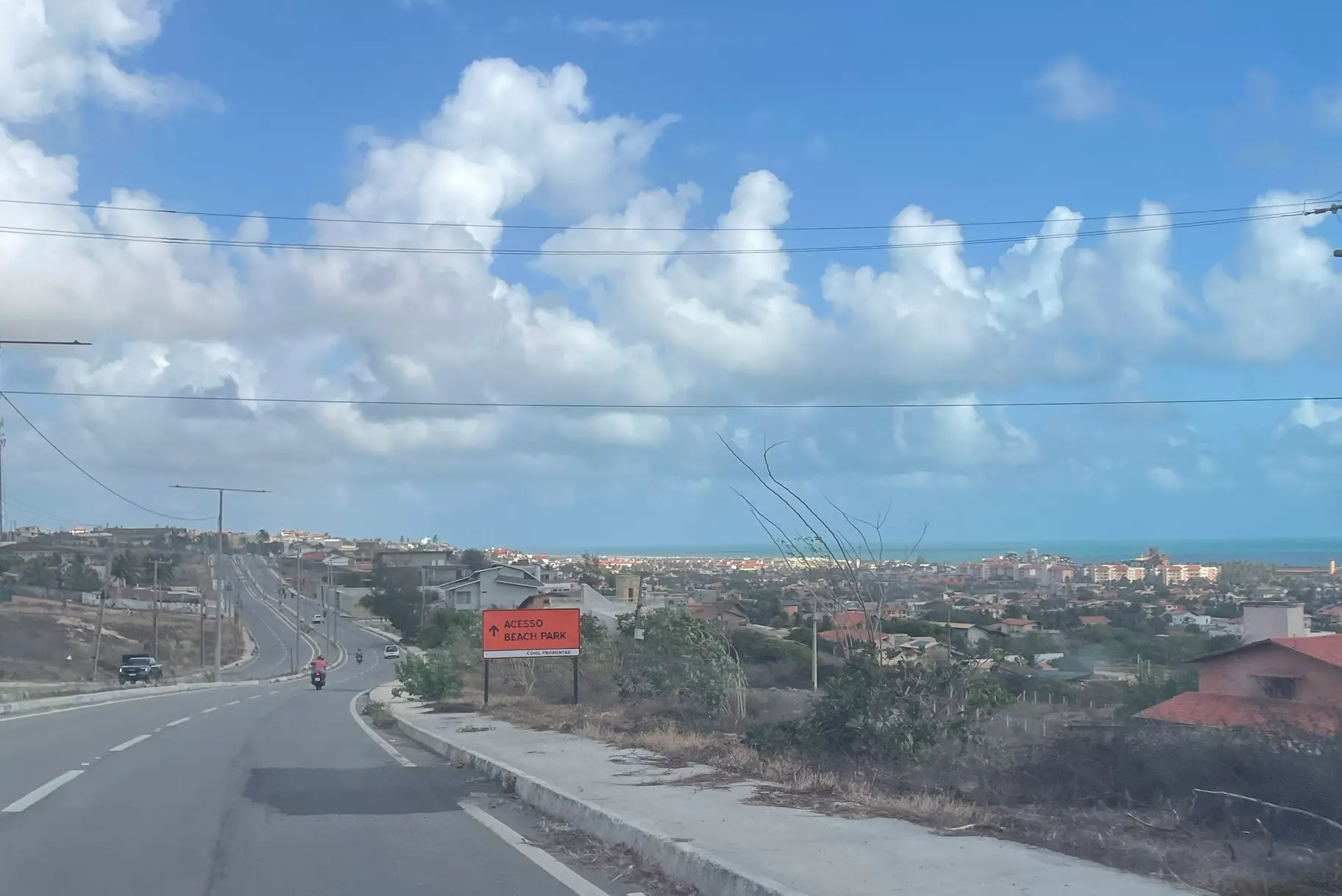 Foto de uma rodovia estadual na cidade de Aquiraz, no Ceará. À direita, há condomínios residenciais e o mar. O céu está bastante nublado com nuvens brancas.