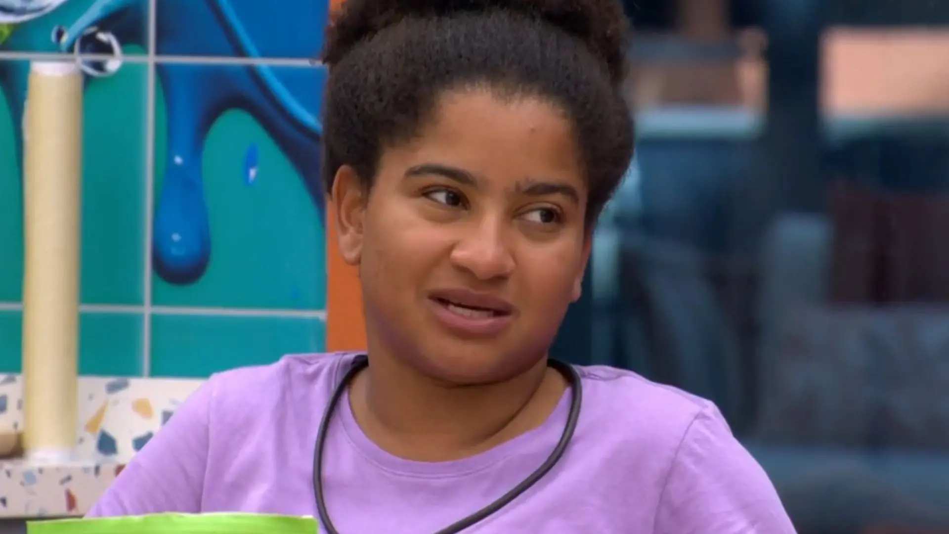 Pessoa sentada à mesa em área interna da casa de reality show, usando camiseta clara, com caneca verde à frente e parede decorada ao fundo.