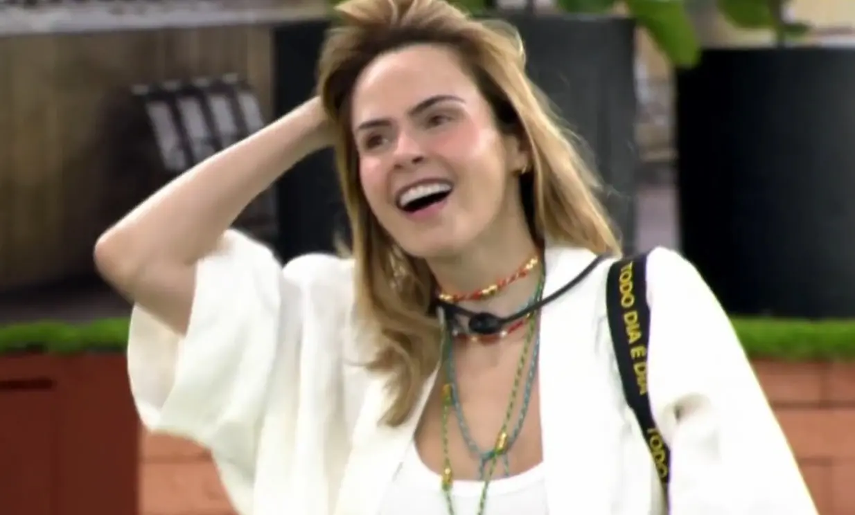 Ana Paula é uma mulher de meia idade, loira, branca, de cabelo liso tamanho médio. Na foto, ela está de branco e mexe no cabelo, sorrindo.